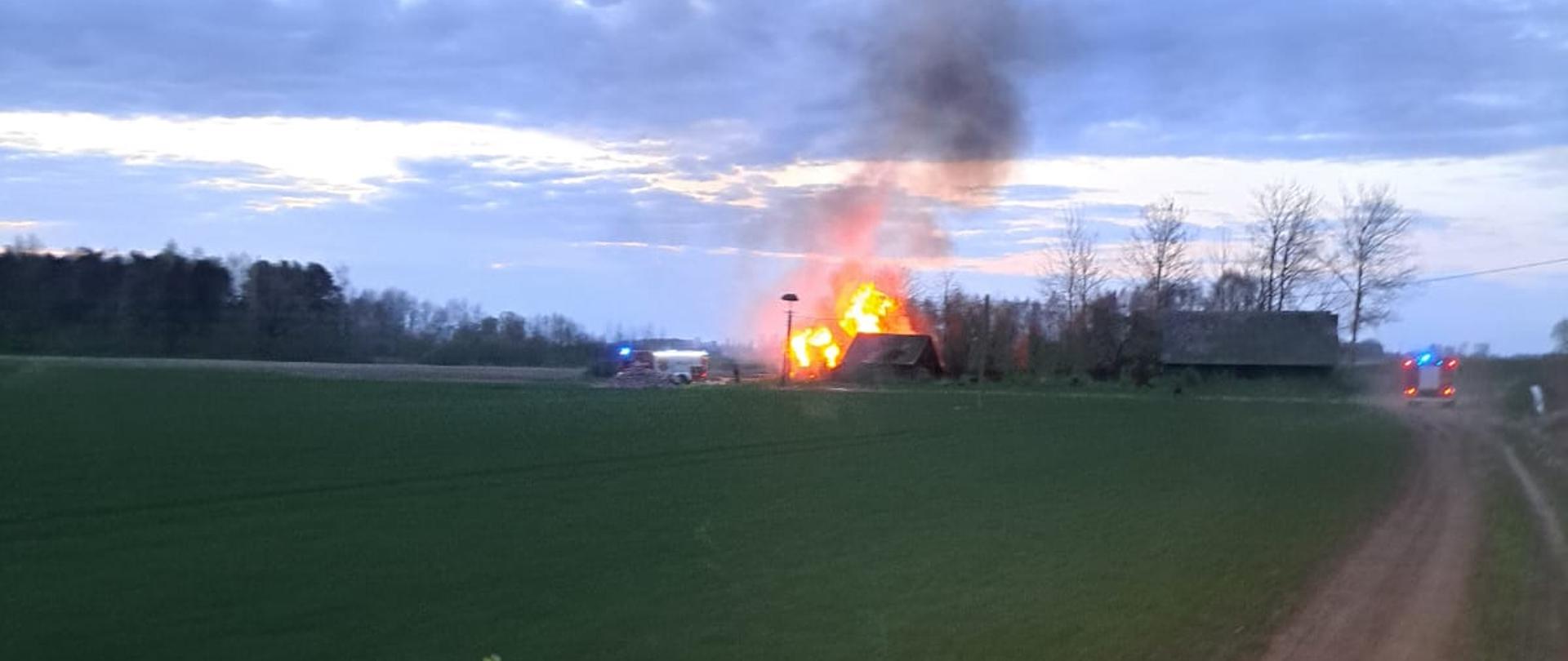 Na polu stoi kilka budynków, jeden z nich się pali. Dojeżdżają do niego strażackie samochody na sygnałach.