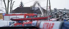 pojazd strazy pożarnej stoi przy budynku, strażacy z kosza znajdującego się na drabinie gaszą palący się dach