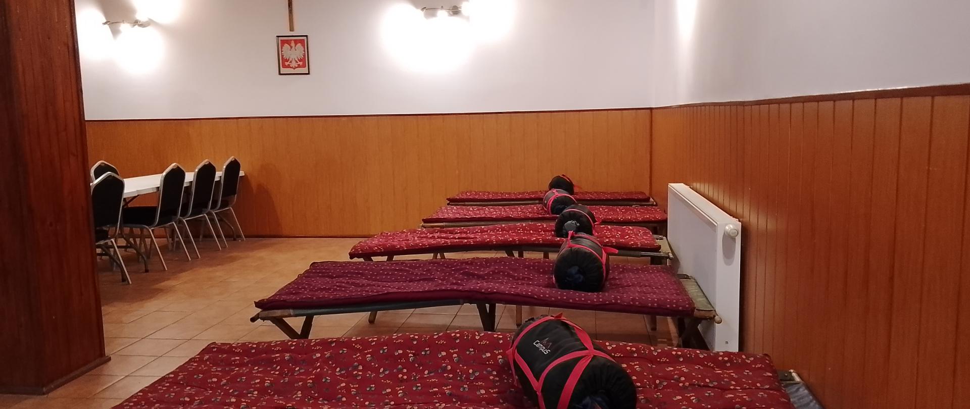 Pomieszczenie z łóżkami polowymi i stołem.