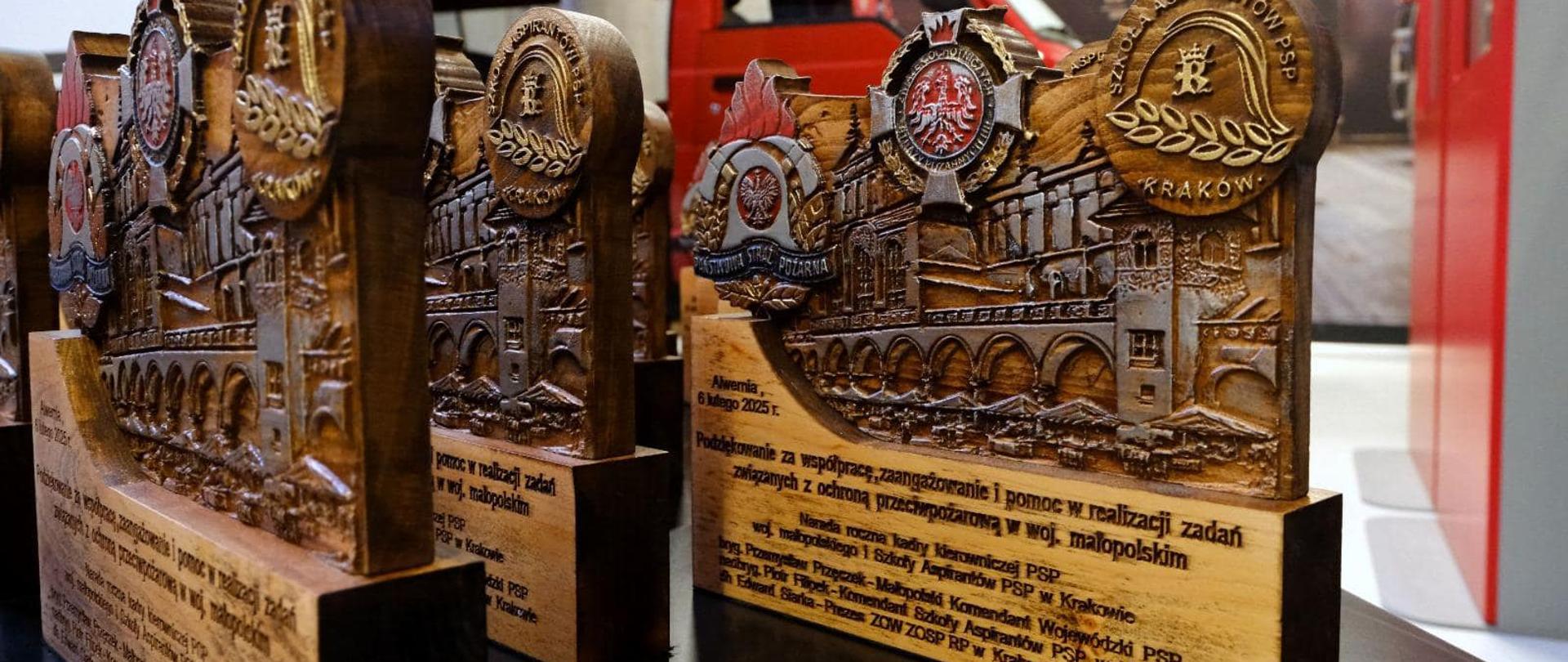 Zdjęcie wykonane w Sali Głównej Małopolskiego Muzeum Pożarnictwa w Alwerni w trakcie narady rocznej. Na zdjęciu widoczne drewniane tabliczki z grawerem, którego tytuł brzmi: „ Podziękowanie za współpracę, zaangażowanie i pomoc w realizacji zadań związanych z ochroną przeciwpożarową w województwie małopolskim” oraz logotypami PSP , SAPSP i ZOSP RP. W tle widać zamazane elementy wystroju auli czerwono – szarą szafę i czerwony samochód pożarniczy, stanowiące eksponaty oraz część ekranu projekcyjnego. 