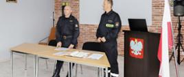 dwóch strażaków ubranych w ubranie koszarowe stoi przed stolikiem, na stole rozłożone dokumenty, w tle ściana z czerwonej cegły na której jest szary ekran projekcyjny, po prawej stronie kadru widoczna mównica z białym orłem na czerwonym tle oraz flaga państwowa stojąca w stojaku, widoczna w tle kolumna głośnikowa barwy czarnej na czarnym stojaku, po lewej stronie zdjęcia wizerunek Matki Boskiej w ramce, parkiet na podłodze w kolorze szarości