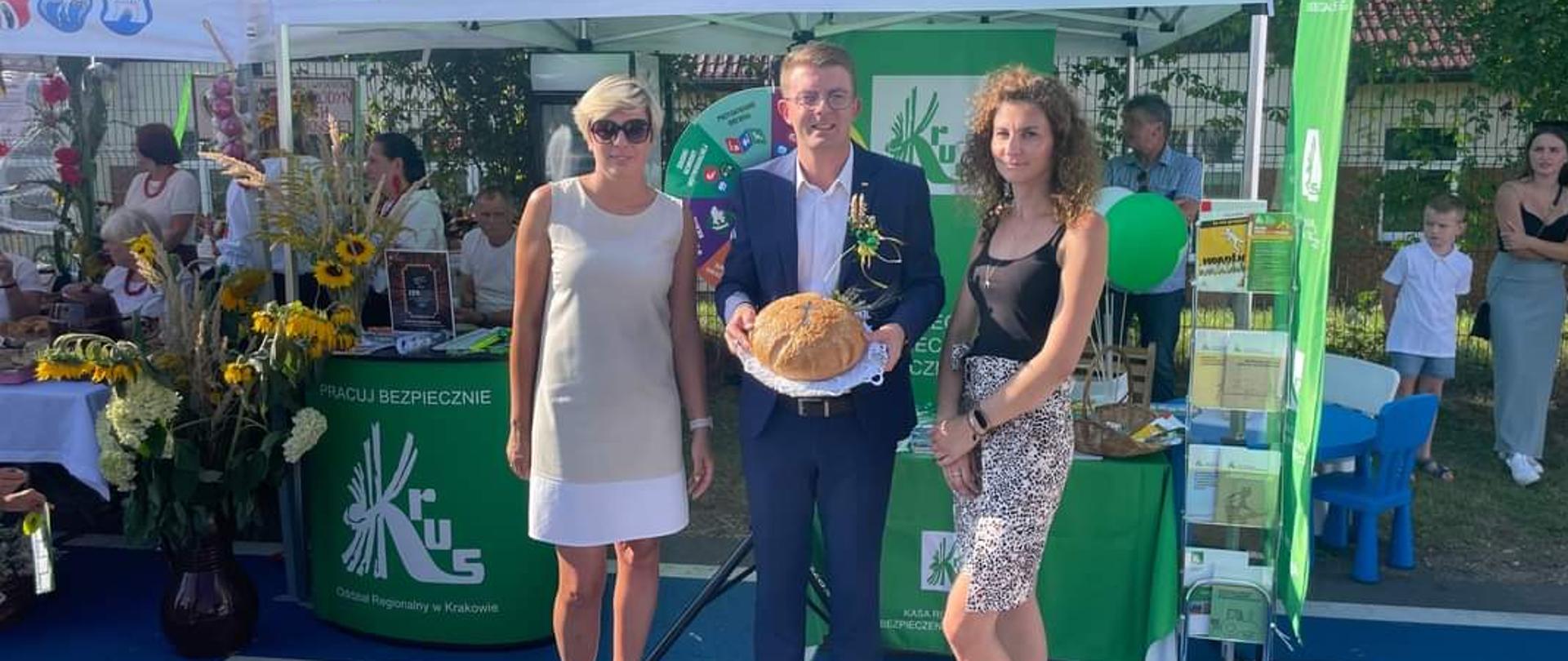 Na tle stoiska informacyjnego KRUS stoją dwie kobiety oraz mężczyzna, który trzyma w rękach bochen chleba.