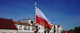Na maszcie powiewa flaga biało-czerwona. W tle budynki.