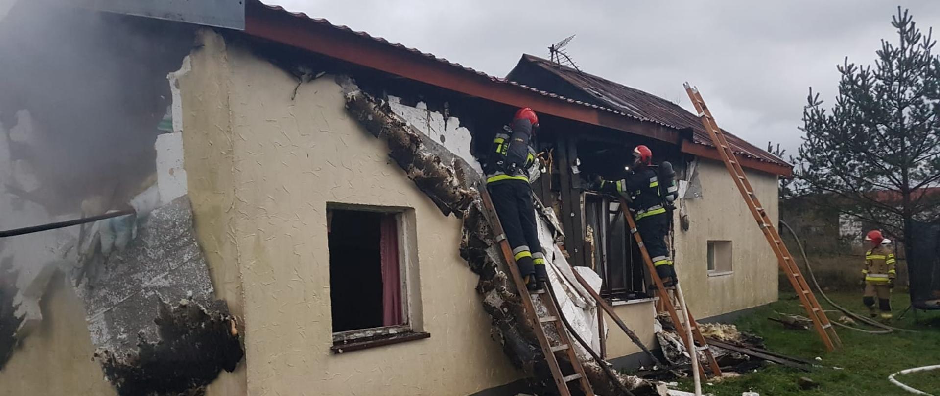 Na zdjęciu widać nadpalony budynek mieszkalny. Z lewej strony widać wypaloną kalenicę budynku. Przy ścianie budynku widać dwie przystawione drabiny na których widać pracujących strażaków rozbierających część ściany w celu dogaszenia ognia.
