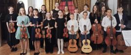Zdjęcie kolorowe. Odświętnie ubrani uczniowie z instrumentami: skrzypcami, wiolonczelami, gitarami pozują do zdjęcia. W tle wysokie okno ze szprosami i fantazyjnie upięte zasłony z lambrekinem w kolorze brązowym.