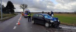 Na zdjęciu znajduje się z rozbitym przodem samochód koloru czarnego. Obok stoją dwaj policjanci u brani w mundury koloru granatowego z białą czapką na głowie. Samochód stoi na jezdni. W tle stoi karetka pogotowia oraz strażacy.