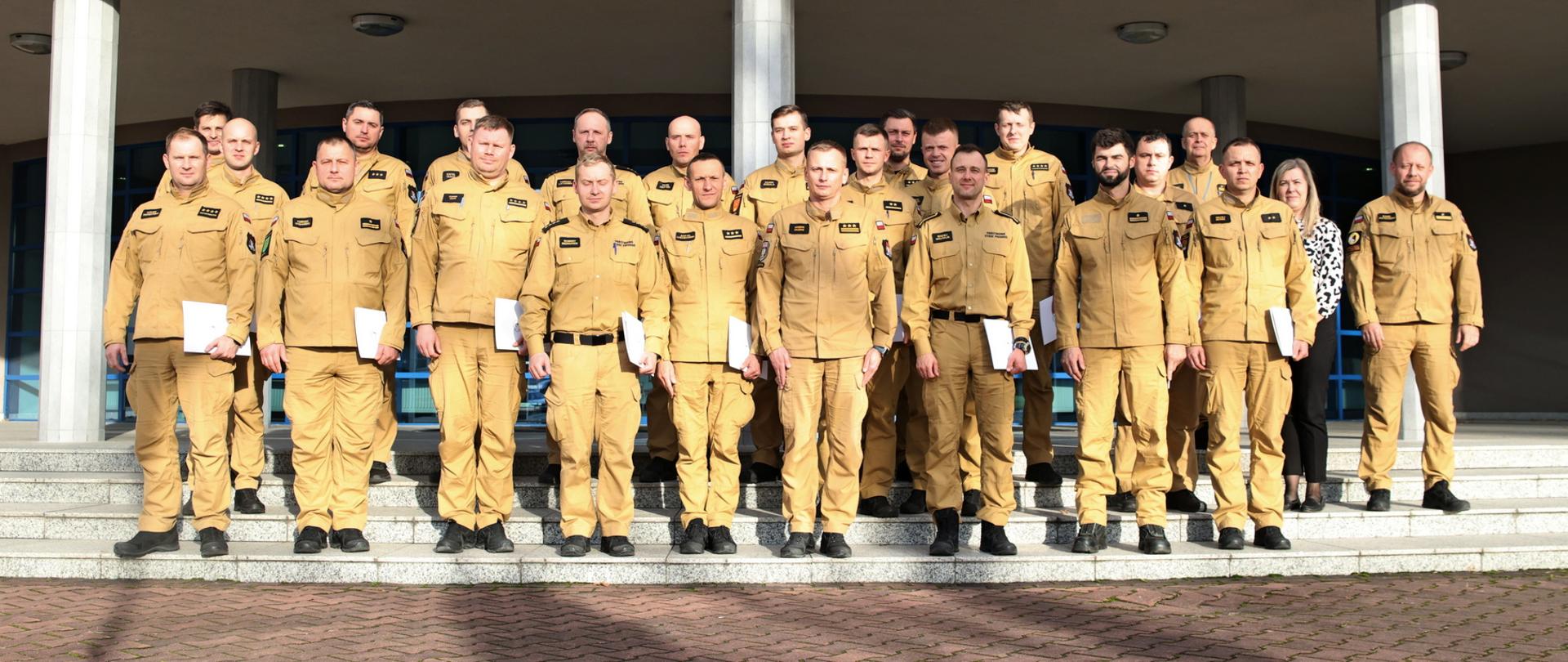 Pamiątkowe zdjęcie grupowe funkcjonariuszy PSP będący uczestnikami szkolenia z zakresu kierowania działaniami ratowniczymi na poziomie taktycznym (20 osób) w towarzystwie Zastępcy Komendanta Centralnej Szkoły PSP, kobiety oraz dwóch funkcjonariuszy CS PSP
