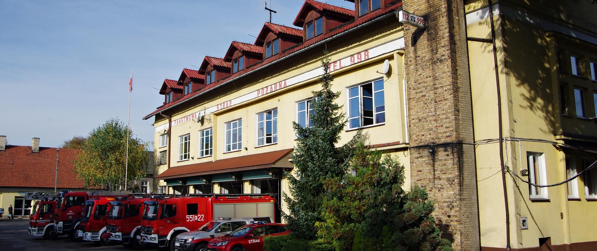 Samochody pożarnicze różnego typu stojące na placu przed budynkiem Jednostki Ratowniczo- Gaśniczej w Wadowicach. Po prawej stronie żółty, trzykondygnacyjny budynek jednostki z czterema bramami garażowymi. Na budynku poniżej dachu na białym pasie czerwony napis Państwowa Straż Pożarna tel. 998.