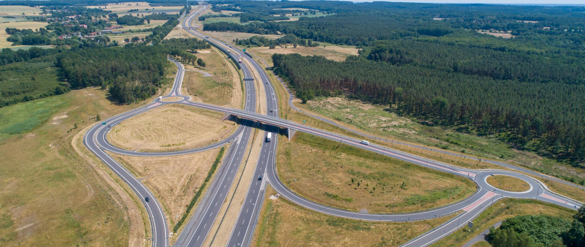 Zdjęcie lotnicze nowoczesnego węzła drogowego z lotu ptaka, z wielopasmową drogą ekspresową, dwoma rondami połączonymi wiaduktem oraz otaczającymi polami i lasem.