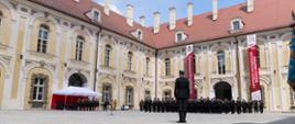 Widok dziedzińca Pałacu Książęcego w Żaganiu podczas uroczystego apelu strażackiego. W oddali widać ustawionych w szyku strażaków oraz dwa pionowe banery informujące o obchodach 30-lecia Krajowego Systemu Ratowniczo-Gaśniczego.