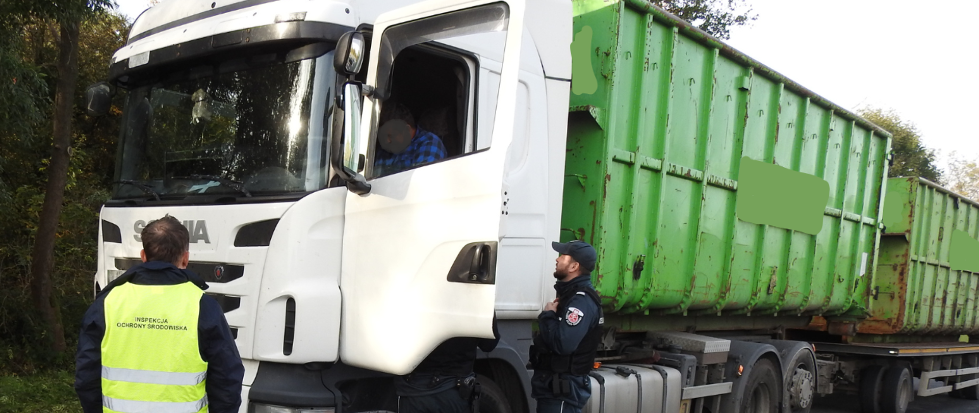 Inspektor Wojewódzkiego Inspektoratu Ochrony Środowiska w Opolu, ubrany w odblaskową kamizelkę z napisem "Inspekcja Ochrony Środowiska". Przed Inspektorem stoi kontrolowany samochód ciężarowy, przy którym stoi dwóch funkcjonariuszy. 