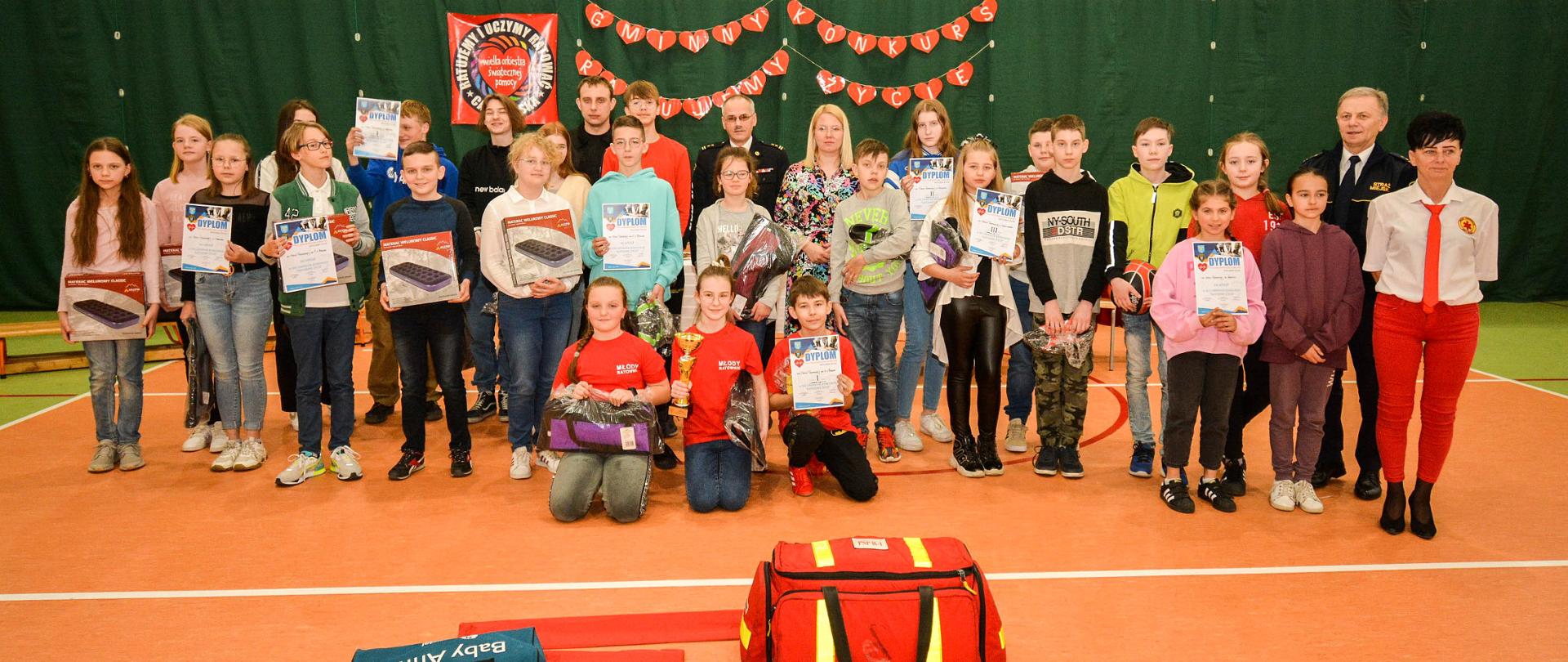 Wspólne zdjęcie wszystkich uczestników konkursu z komisją konkursową. U dołu zdjęcia torba medyczna PSP-R1 i manekin do resuscytacji