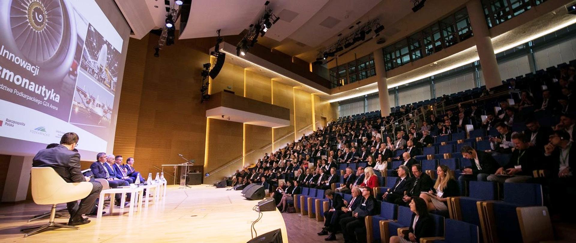 W auli na scenie siedzi przy stolikach sześciu mężczyzn. Na wprost siedzą słuchacze.
