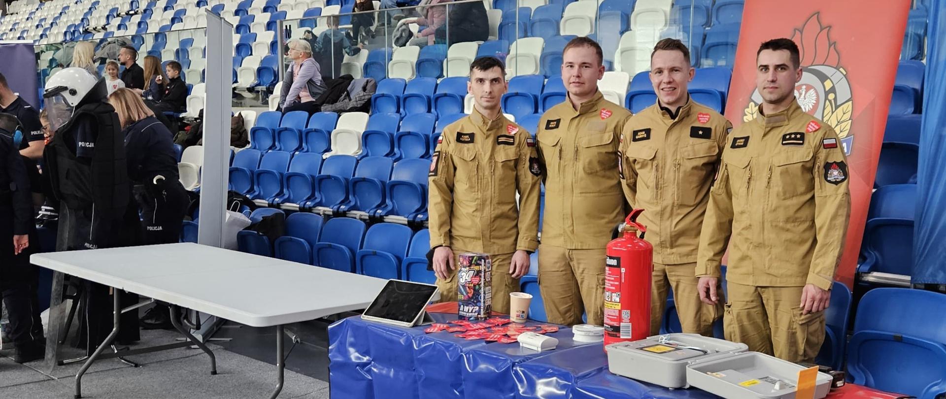Portret grupowy strażaków stojących przy niebieskim stoisku z logo "Firemax Rescue Systems". Na stoisku znajdują się ulotki, puszka na datki WOŚP oraz gaśnica. Obok stoi baner "Państwowa Straż Pożarna – Komenda Powiatowa PSP w Mielcu". W tle widoczne są puste trybuny hali z biało-niebieskimi krzesełkami.
