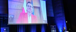 Adriana Porowska, Minister ds. Społeczeństwa Obywatelskiego widoczna na ekranie, gdy zdalnie uczestniczy w konferencji "Społeczeństwo obywatelskie jako czynnik budowania odporności na zjawiska kryzysu we współczesnej Europie. Doświadczenia i wyzwania". Ministra przemawia podczas sesji otwierającej konferencję.