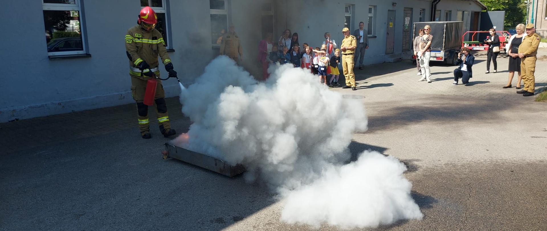 Strażak przy użyciu gaśnicy proszkowej gasi pożar paliwa w tacy. Z tacy unoszą się kłęby szarego dymu. Na drugim planie dzieci wraz z opiekunami oraz towarzyszący im strażacy.