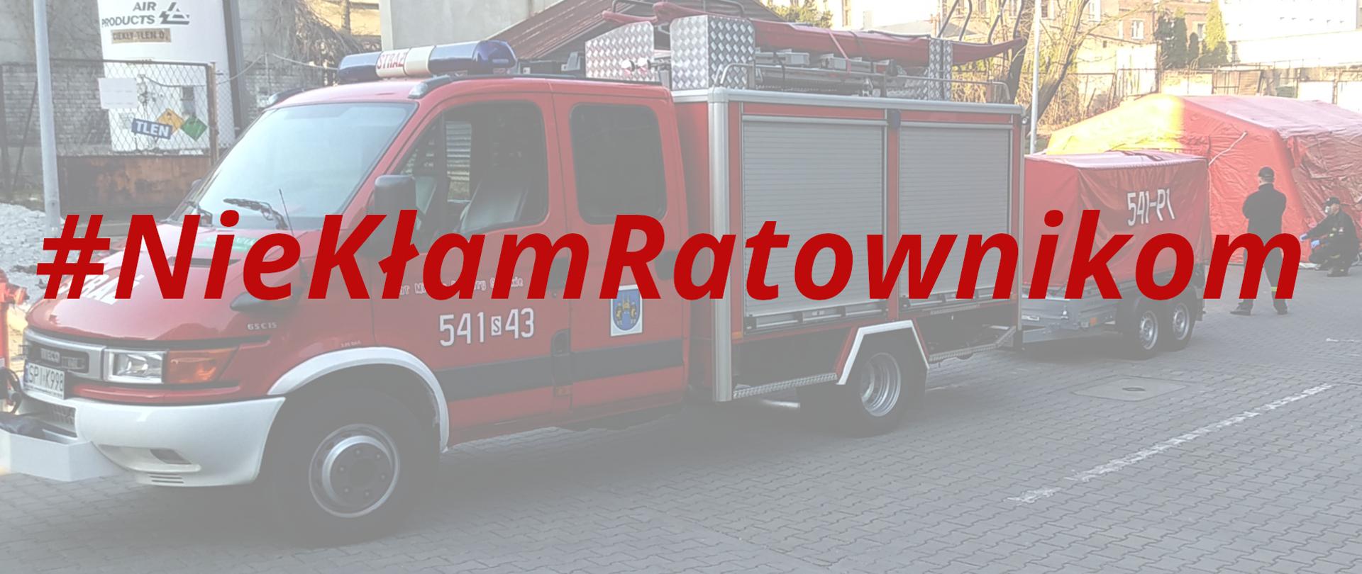 Ilustracja przedstawia duży czerwony napis #NieKłamRatownikom. W tle zamglone zdjęcie lekkiego samochodu ratownictwa technicznego wraz z przyczepą oraz rozstawionego namiotu.