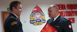 Świetlica komendy. Komendant powiatowy trzymając w lewej ręce teczkę, prawą dłonią ściska dłoń awansowanego strażaka, składając mu przy tym gratulacje. Strażacy mają na sobie mundury wyjściowe. W tle na ścianie znajduje się logo PSP oraz napis: Komenda Powiatowa Państwowej Strazy Pożarnej w Rawiczu.