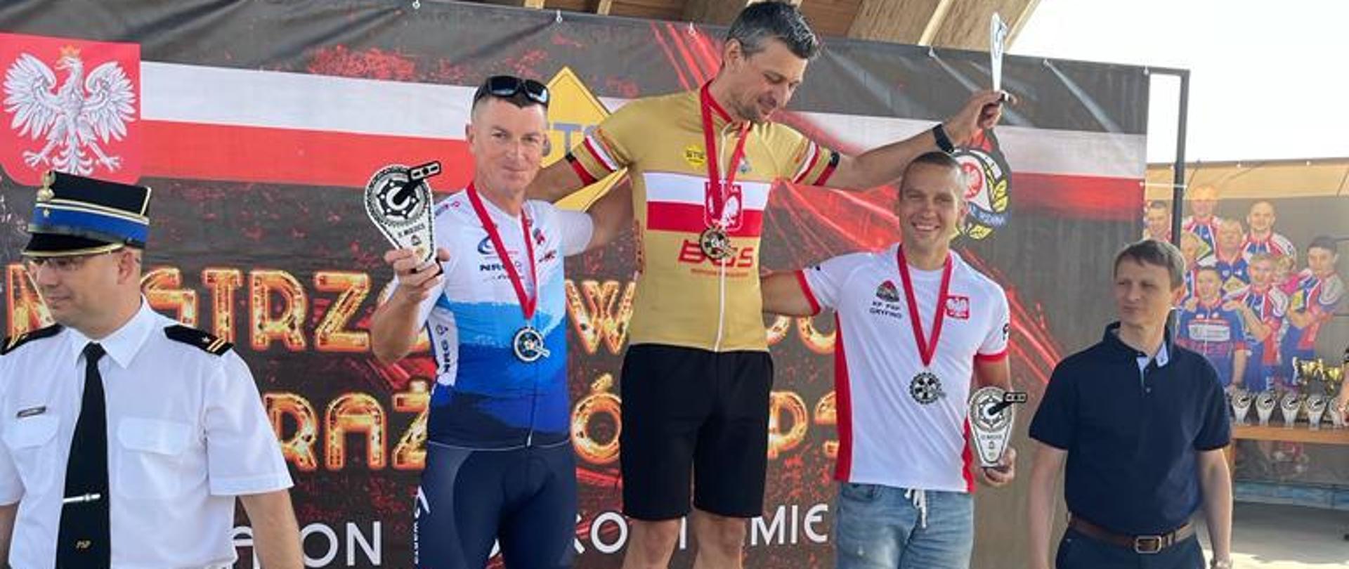 trzech zawodników w strojach kolarskich stojących na podium, z lewej strażak OSP w białej wyjściowej koszuli, po prawej mężczyzna w niebieskiej koszulce polo, w tle baner zawodów kolarskich