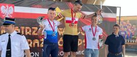 trzech zawodników w strojach kolarskich stojących na podium, z lewej strażak OSP w białej wyjściowej koszuli, po prawej mężczyzna w niebieskiej koszulce polo, w tle baner zawodów kolarskich