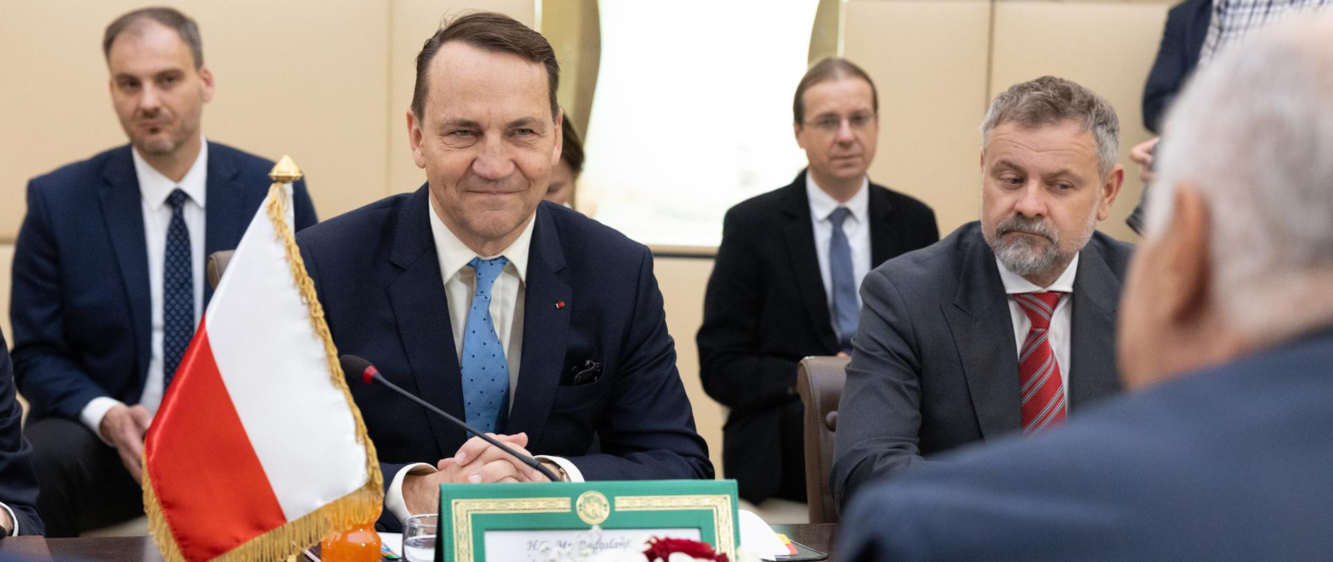 Na pierwszym planie Wicepremier Radosław Sikorski w granatowym garniturze i jasnoniebieskim krawacie. Na stole widoczna jest flaga Polski, mikrofon, szklanki z wodą oraz dokumenty, a na pierwszym planie ktoś gestykuluje, siedząc tyłem do kamery.