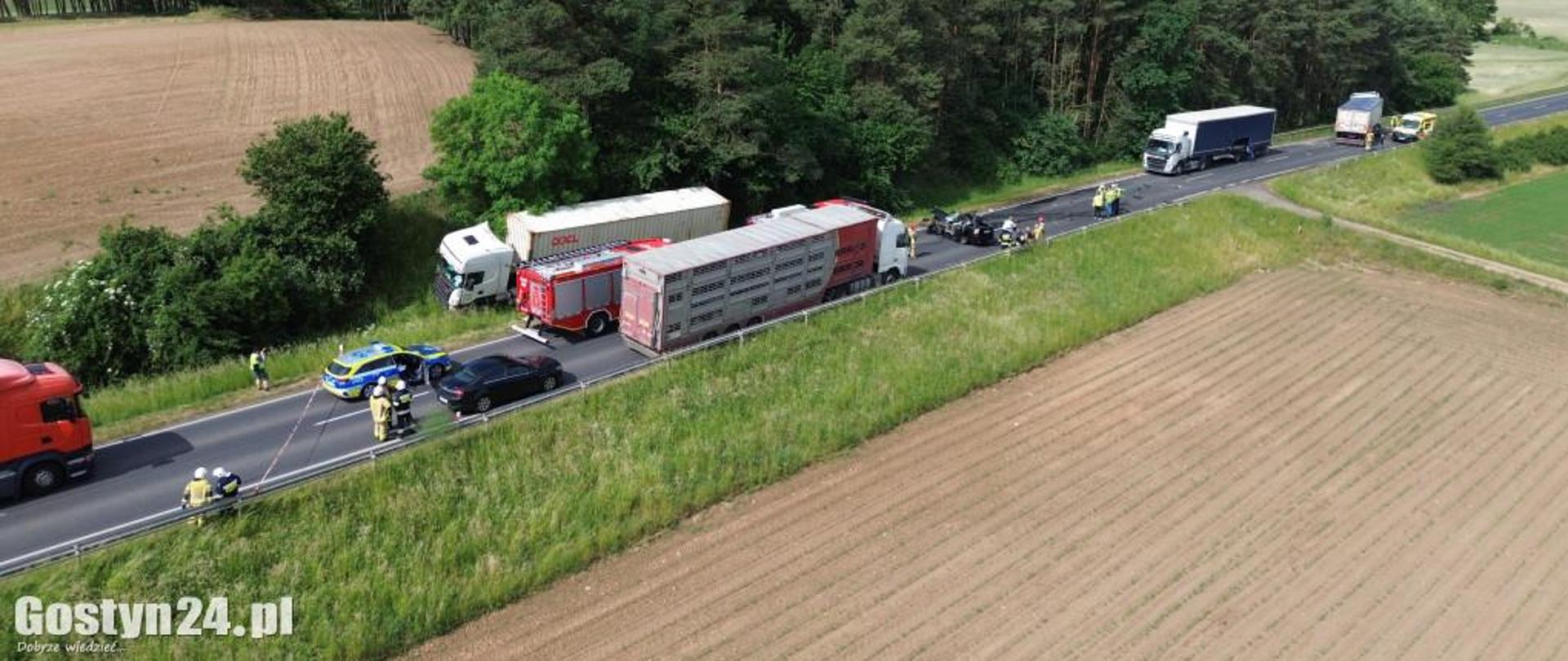 Zdjęcie z lotu ptaka. Przedstawia zastępy straży pożarnej, uszkodzone pojazdy oraz ratowników. Wszyscy znajduję się na drodze. W tle jest mały las oraz pola.