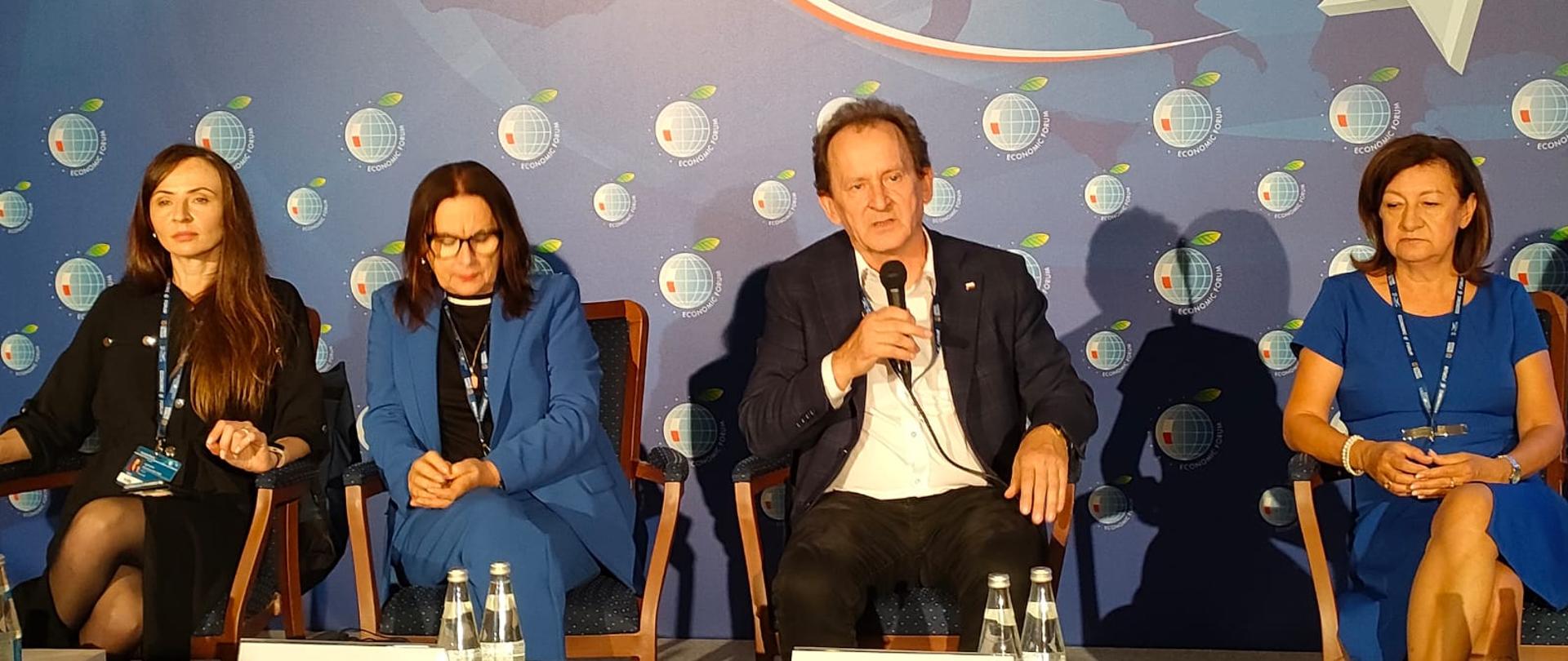 Wiceminister Bernacki siedzi na krześle obok kilku osób, mówi do trzymanego w ręku mikrofonu, za nimi niebieska ściana.