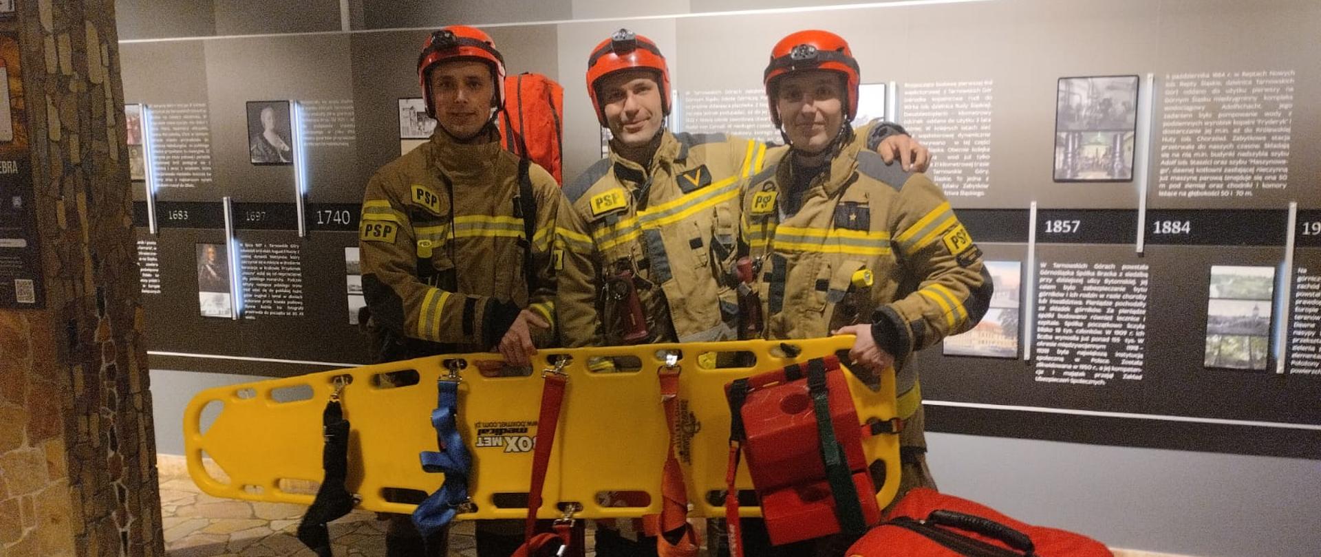 Trzech strażaków stoi w mundurach bojowych. W rękach trzymają żółtą deskę ortopedyczną.