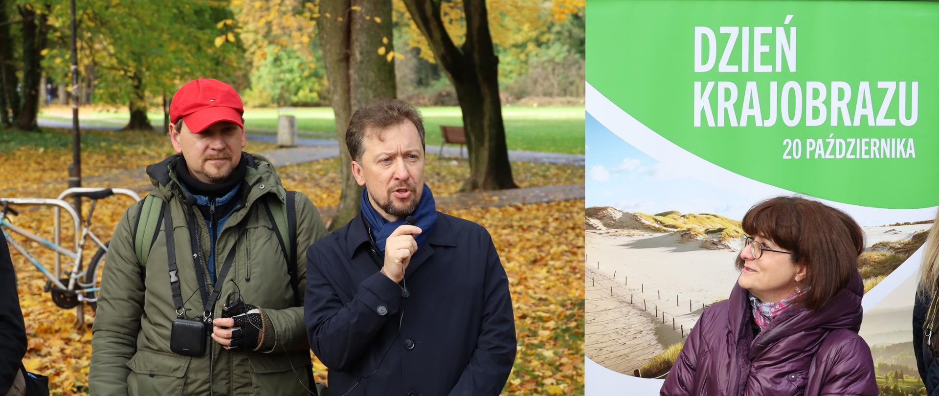 Na tle jesiennego parku (drzewa, pożółkłe liście) stoi dwóch mężczyzn i kobieta. Jeden z mężczyzn trzyma mikrofon, z za plecami kobiety plakat z napisem: Dzień Krajobrazu, 20 października.
