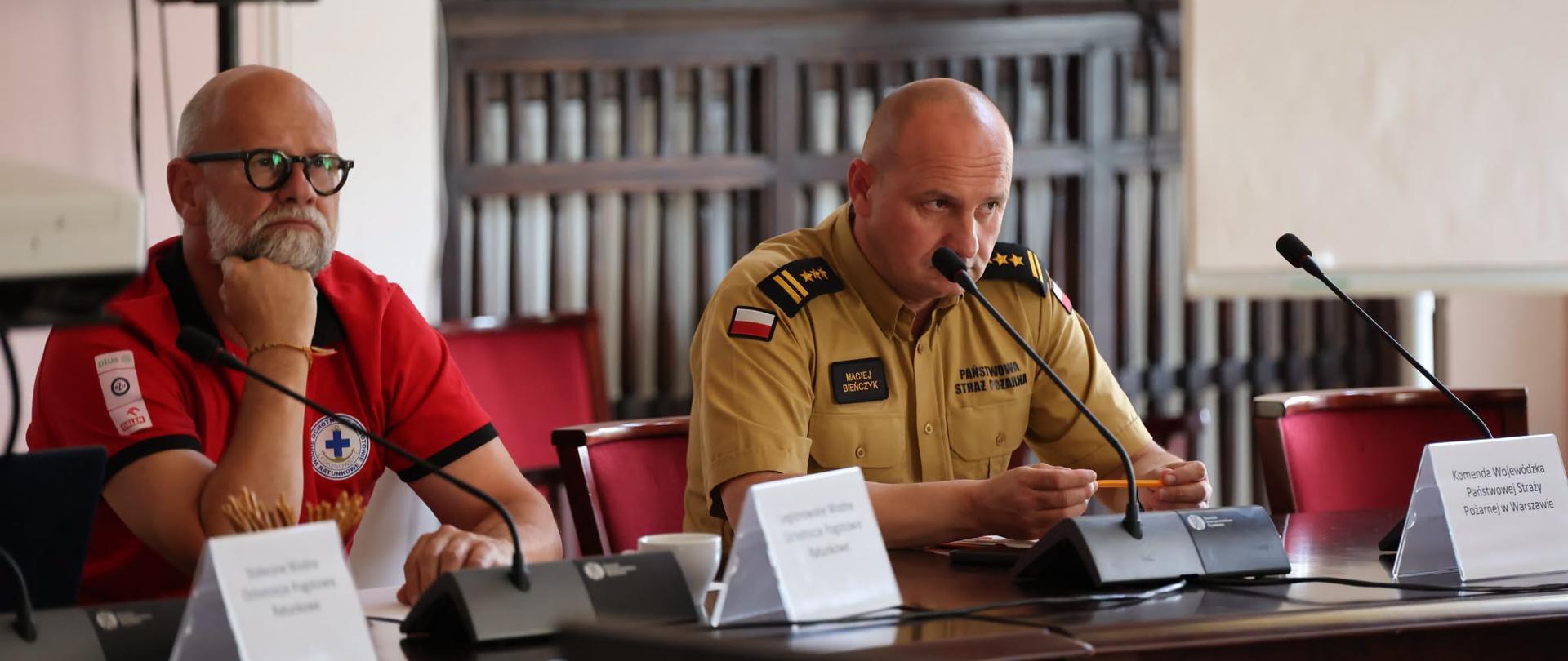 Na zdjęciu przedstawiono dwóch uczestników spotkania siedzących przy stole konferencyjnym. Po lewej stronie znajduje się mężczyzna w czerwonej koszulce, a po prawej funkcjonariusz PSP w mundurze służbowym koloru piaskowego. Na stole stoją mikrofony, dokumenty i tabliczki z nazwiskami. W tle widoczne są ściany sali konferencyjnej i sprzęt nagłośnieniowy.