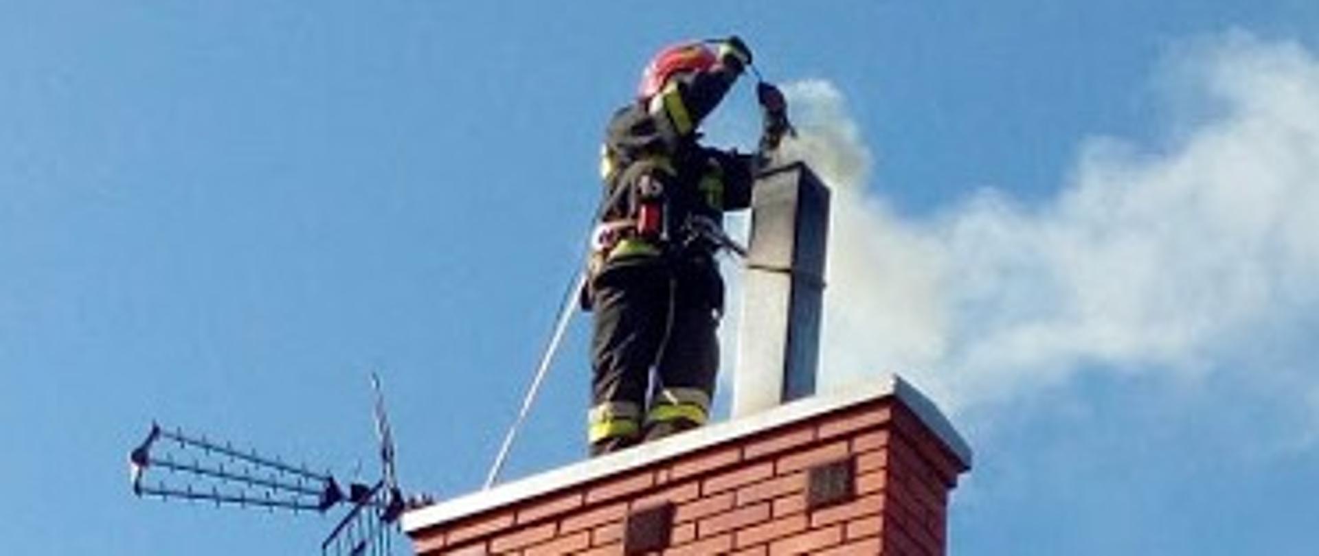 Zdjęcie przedstawia strażaka stojącego na kominie, który gasi pożar sadzy. W tle błękitne niebo.