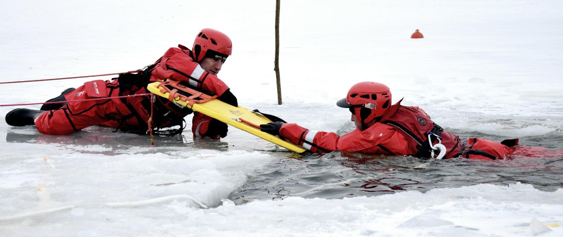 Zdjęcie zrobione na zewnątrz, nad zamarzniętym zbiornikiem wodnym. Dwóch strażaków, ubranych w skafandry zabezpieczające przed wychłodzeniem. Ratownicy ćwiczą działania na zamarzniętym akwenie. Jeden z nich (po prawej stronie zdjęcia) zanurzony jest w wyciętym w lodzie przeręblu. Drugi strażak leżący na lodzie, używając deski ortopedycznej próbuje wyciągnąć strażaka z przerębli na lód.