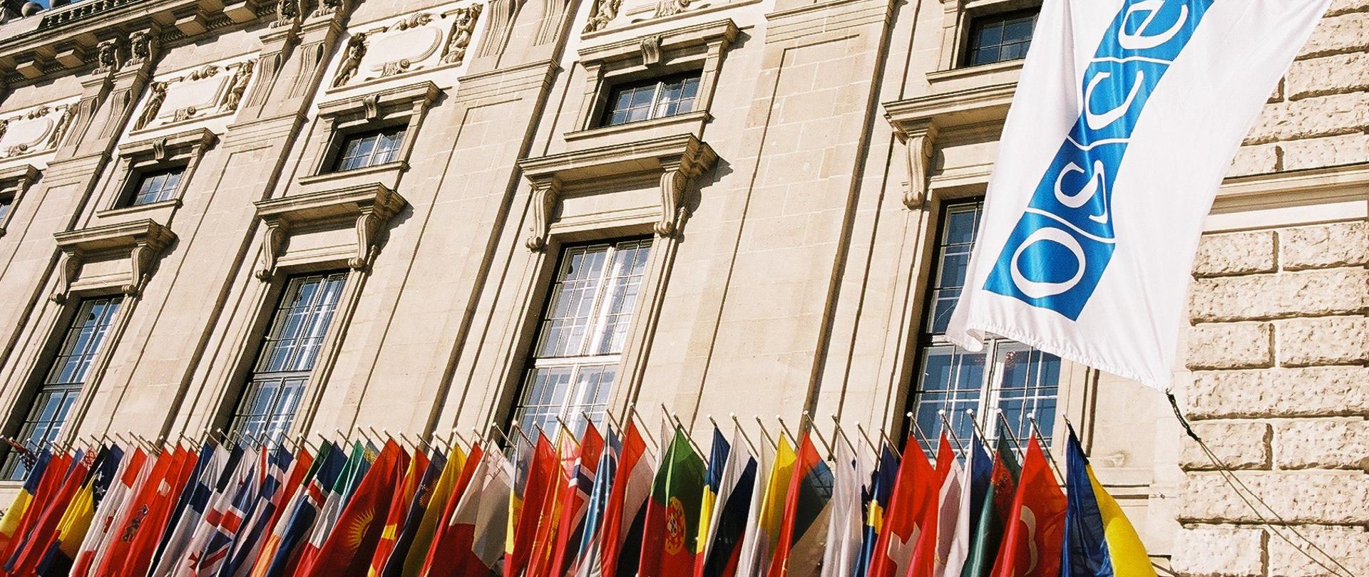 flaga OBWE oraz flagi 57 państw uczestniczących OBWE na tle Pałacu Hofburg w Wiedniu