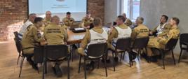 Strażaków, którzy siedzą przy jednym stole i omawiają zakres ćwiczeń. Kilku z nich ma założone kamizelki osób funkcyjnych w sztabie. Za nim w tle, na rzutniku prezentowane są zagadnienia.