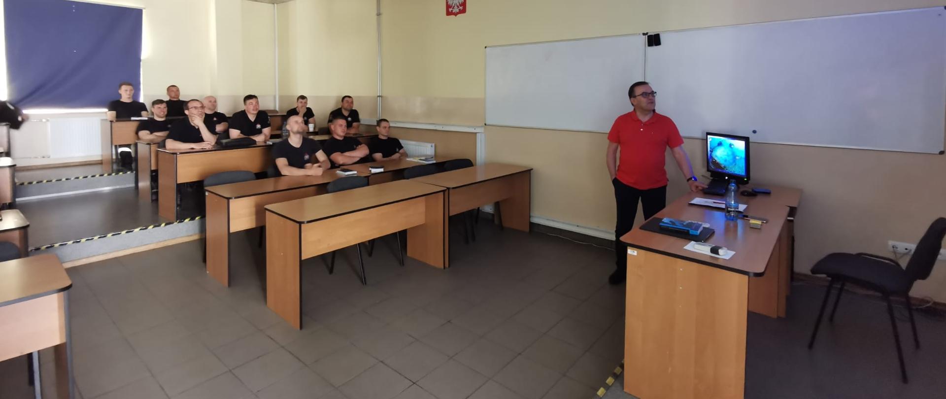 Na sali wykładowej za biurkami siedzi 27 słuchaczy w trakcie szkolenia po prawej stornie widać prowadzącego szkolenie 