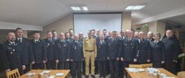 Strażacy Państwowej Straży Pożarnej oraz ochotniczych straży pożarnych stoją obok siebie przed stołami na których rozłożony jest poczęstunek i leżą dokumenty.