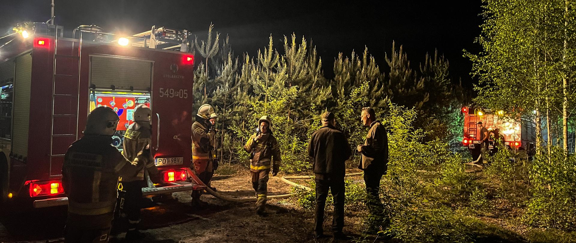 Na zdjęciu strażacy w ubraniach specjalnych stoją przy samochodzie strażackim po lewej stronie, który oświetla teren działań. Na środku stoją leśnicy i obserwują działania gaśnicze. W tle widać las. Po prawej stronie w głębi lasu stoi samochód gaśniczy. Zdjęcie wykonane porą nocną.