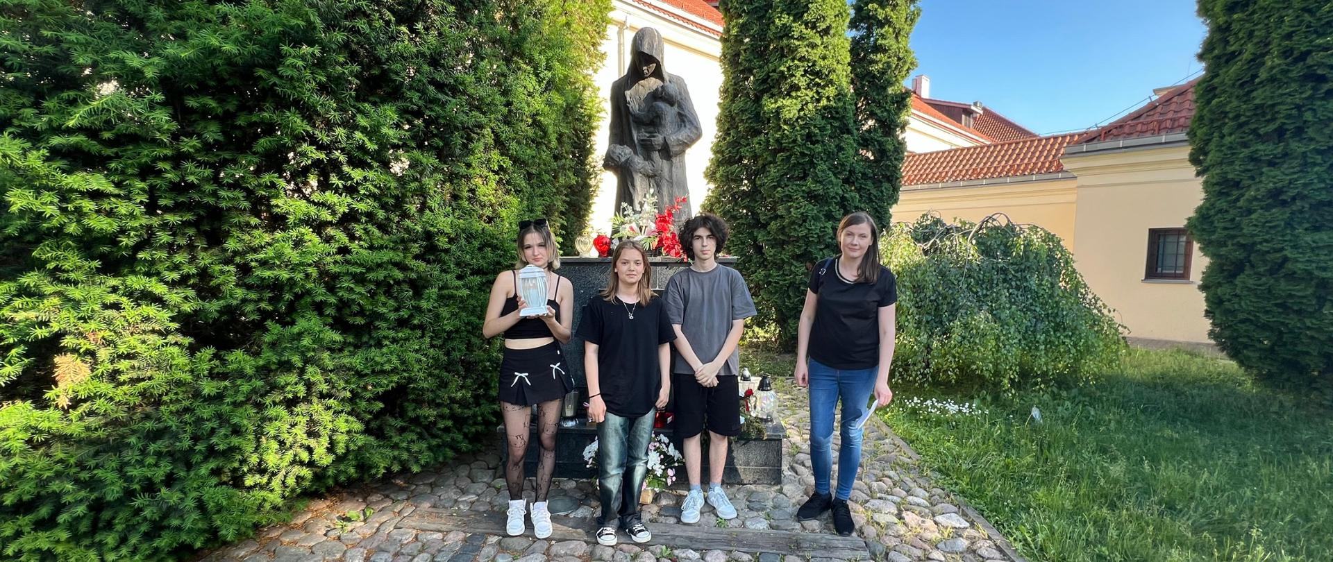 Po pomnikiem "Matkom Sybiraczkom" stoi troje wychowanków i wychowawczyni, dziewczyna stojąca z lewej strony trzema biały znicz. Pomnik jest udekorowany kwiatami białymi i czerwonymi, na cokole stoją znicze. w tle widać fragmenty budynku z czerwonym dachem, wokół zielone drzewa i kwiaty, z przodu wybrukowana ścieżka