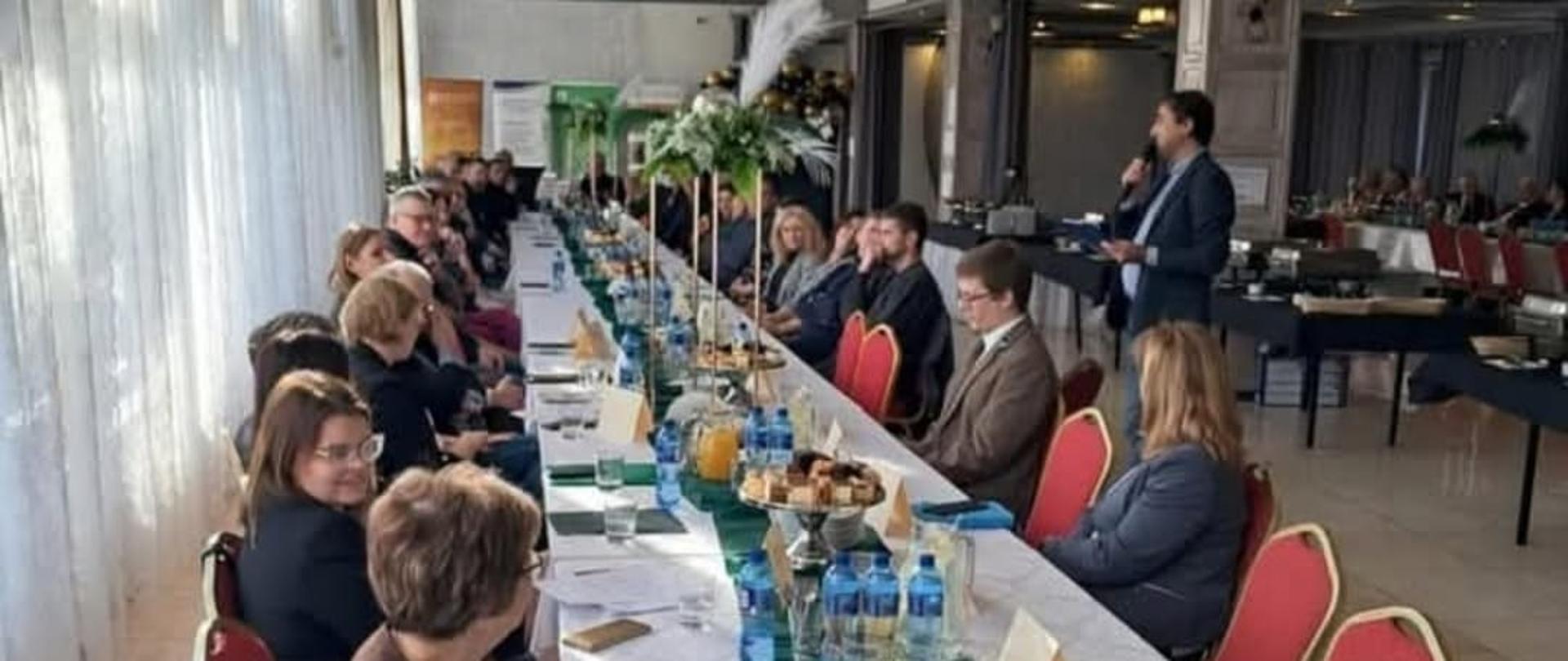 Uczestnicy forum siedzący przy długim stole, przy którym przemawia do mikrofonu przedstawiciel Oddziału w Górze 