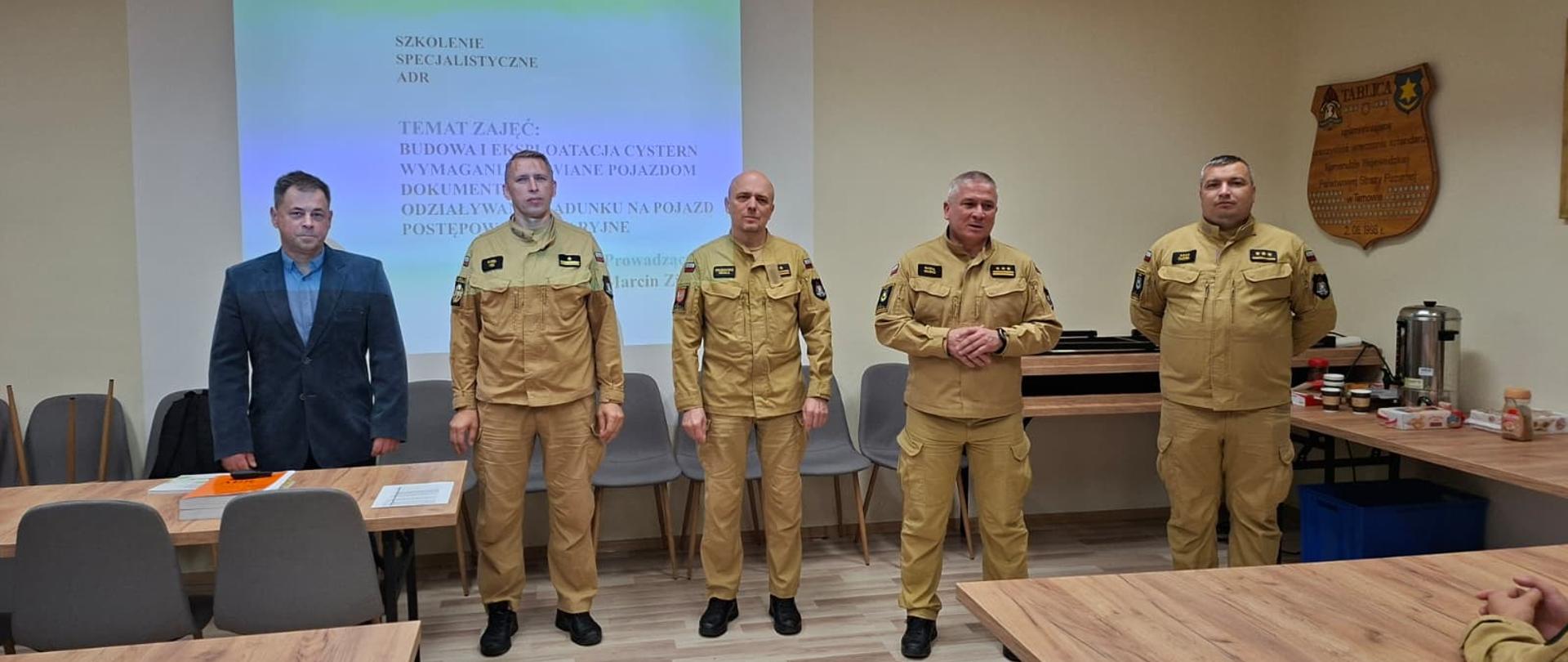 Strażacy stoją w rzędzie mają ubrane piaskowe mundury, obok nich stoi pan w garniturze. Z tyłu widać wyświetloną projekcje na ścianie. Wszystkie osoby znajdują się na sali w której rozmieszczone są ławki.