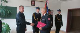 Komendant Powiatowy PSP wręcza akt mianowania nowo przyjętemu funkcjonariuszowi, w tle widzimy poczet sztandarowy
