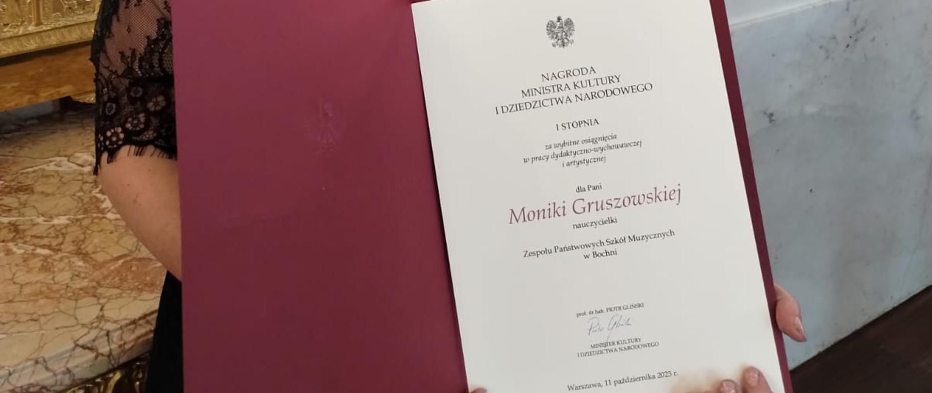 Kobieta w pozycji stojącej trzymająca w dłoniach dyplom z nagrodą. W tle marmurowe kolumny i lustro z Sali Zamku Królewskiego w Warszawie.