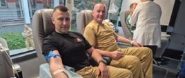 nętrze mobilnego punktu krwiodawstwa. Na pierwszym planie mężczyzna siedzi w fotelu i oddaje krew – do jego przedramienia podłączona jest aparatura medyczna, a obok znajduje się pojemnik z pobieraną krwią. Obok siedzi drugi mężczyzna, również przygotowany do oddania krwi. W tle personel medyczny obsługuje kolejnego dawcę. Przez okna widać budynek i osoby znajdujące się na zewnątrz. Atmosfera jest spokojna i zorganizowana, charakterystyczna dla akcji krwiodawstwa.