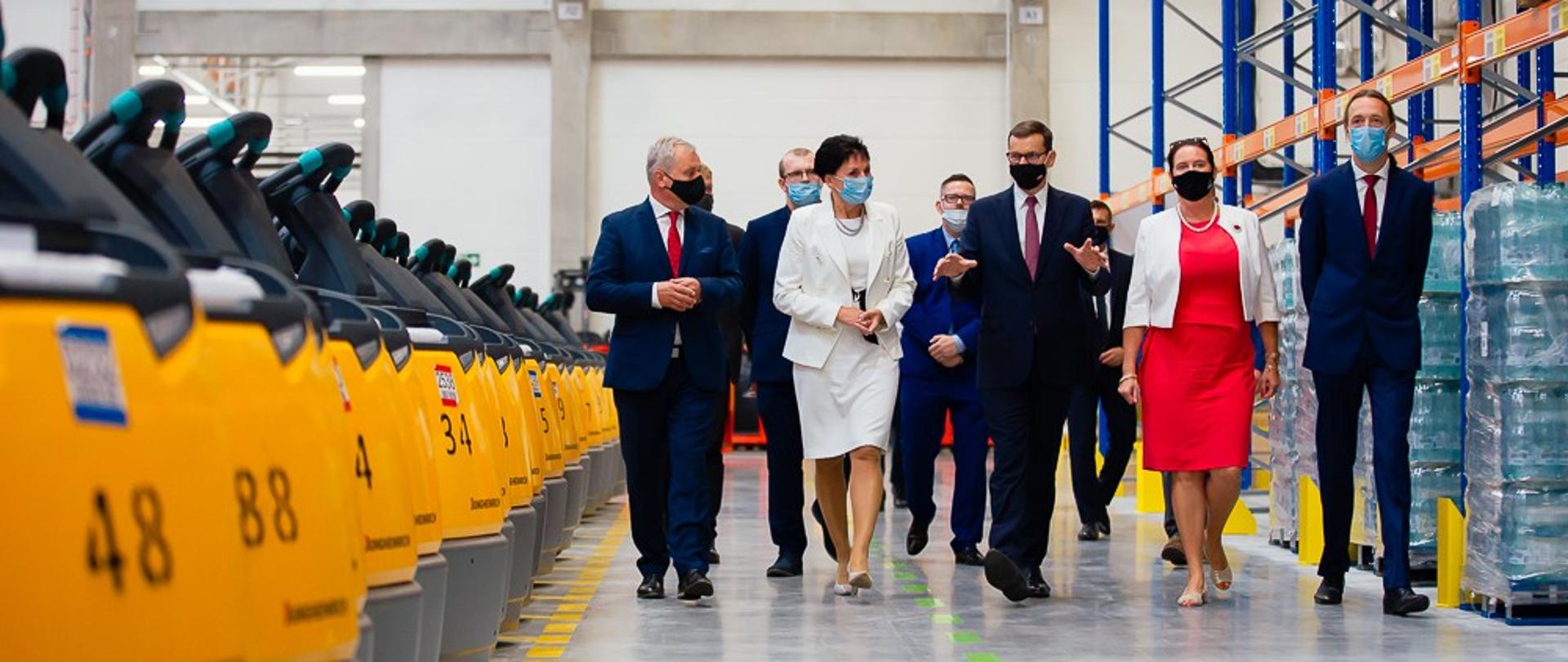 Na zdjęciu 8 osób, idąca w szeregu. W tle magazyn. Po lewej stronie kadru w rzędzie stoją wózki widłowe, po prawej stronie stelaże metalowe. Wśród osób idących w szeregu, w środku, idzie premier. Premier ubrany jest w ciemny garnitury, na twarzy ma czarną maseczkę. Ma otwarte dłonie. 