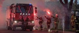 Zdjęcie przedstawia szpaler strażaków w ubraniach specjalnych, którzy trzymają w ręku race z wydobywającym się płomieniem i dymem koloru czerwonego. Środkiem drogi jedzie nowy wóz bojowy. 