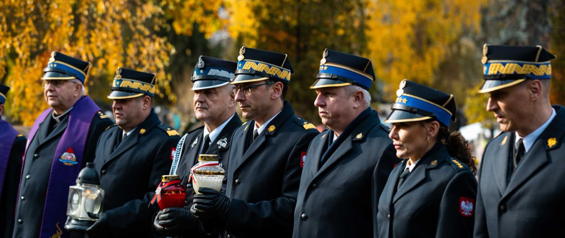 Grupa oficerów PSP i kapelanów stoi w szeregu podczas uroczystości na cmentarzu, trzymając znicze w barwach narodowych; w tle drzewa w jesiennych kolorach.