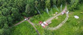 Widok z góry, na leśnej polanie widoczne samochody gaśnicze ustawione przy rozkładanym zbiorniku, strażacy z liniami gaśniczymi podają prądy wody na drzewa. 