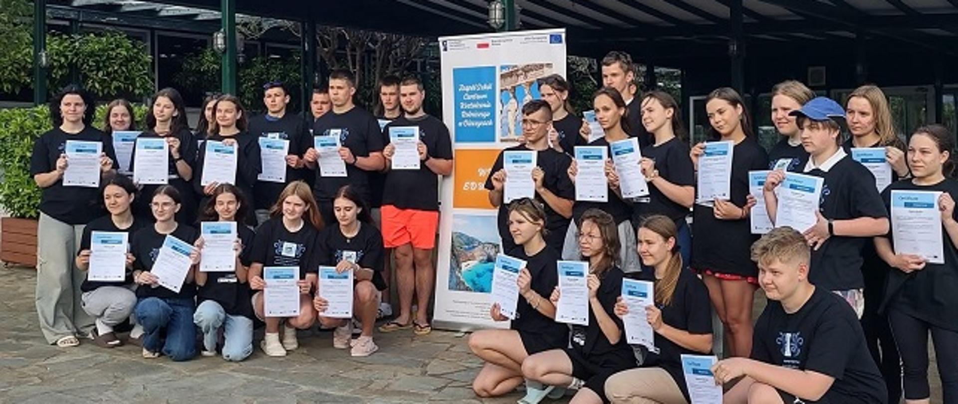Uczniowie pozują do zdjęcia z certyfikatami, stojąc pod wiatą przy roll-up zawierającym opis projektu. 