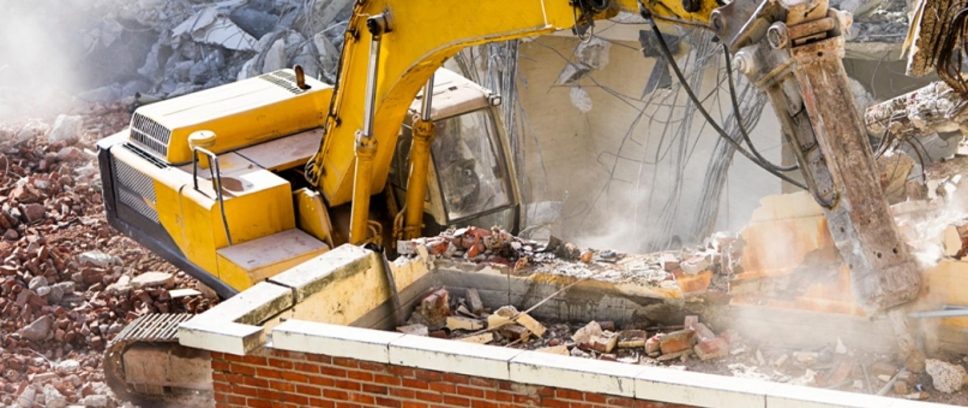 Żółta koparka w trakcie wyburzania budynku; wokół gruz, cegły i kurz unoszący się w powietrzu.
