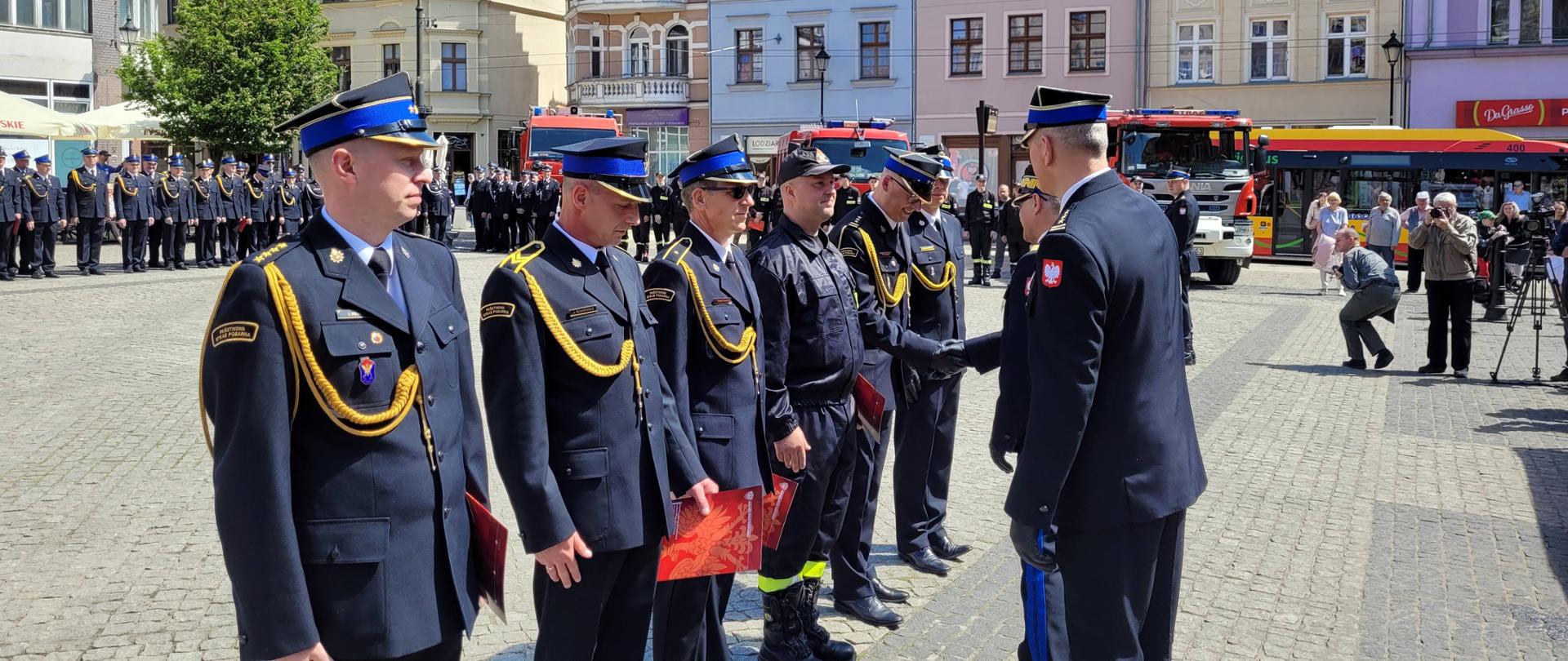 Moment dekorowania strażaków stojących w szeregu przez innych strażaków. W tle zbiórka strażaków, czerwone samochody i zabytkowe kamienice.